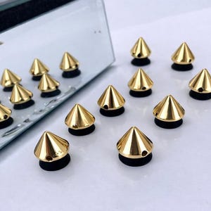 May include: Gold-colored, cone-shaped metal studs with a pointed top and a black base. The studs are arranged on a white surface, reflecting in a mirror. These decorative studs are ideal for adding a unique touch to clothing, accessories, or other craft projects.