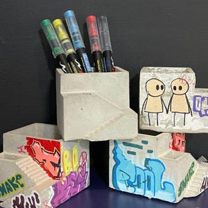 以下が含まれることがあります： 落書きアートが施されたコンクリート製のペン立て。1つのホルダーには、緑、黄、青、赤のインクが入ったペンが入っています。他のホルダーには、人物や文字など、カラフルな落書きデザインが施されています。ホルダーは暗い面に置かれています。
