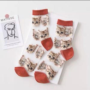 May include: Sheer white socks featuring a repeating pattern of cat faces in brown and tan. The socks have rust-coloured heels and cuffs. A print of a Matisse drawing is in the background.