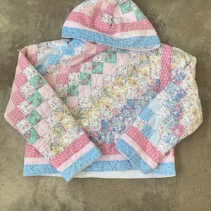 May include: A hooded quilted jacket in pastel shades of pink, blue, and floral patterns. The patchwork design is complemented by long sleeves and a white lining, creating a vintage aesthetic.