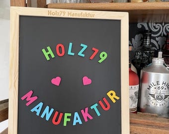 Puede incluir: Una pizarra negra con marco de madera, que presenta el texto "Holz79 Manufaktur" y una cara sonriente hecha con letras magnéticas de colores. Las letras deletrean "HOLZ79" y "MANUFAKTUR". La dirección del sitio web "www.holz79.de" está impresa en la parte inferior de la pizarra.