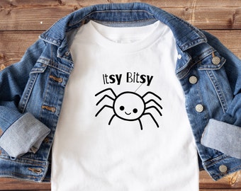 May include: White t-shirt with a black and white cartoon spider and the text "Itsy Bitsy" printed on it. The t-shirt is laying on a wooden surface with a denim jacket draped over it.