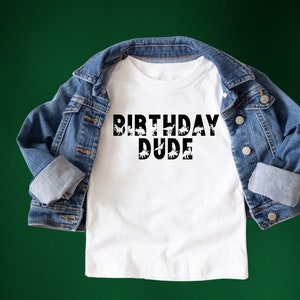 Puede incluir: Camiseta blanca con texto negro que dice "Birthday Dude" con siluetas de dinosaurios dentro de las letras.