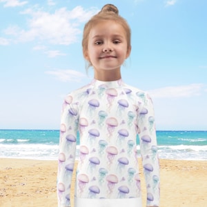 Può includere: Una rash guard bianca a maniche lunghe con una stampa di meduse ad acquerello colorate. La rash guard ha un collo alto e maniche lunghe.