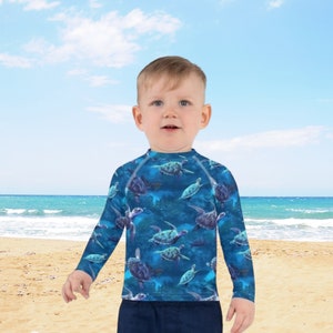 Può includere: Un bambino piccolo che indossa una rash guard a maniche lunghe blu con una stampa di tartarughe marine. La rash guard ha un collo alto e maniche lunghe. Il bambino è in piedi su una spiaggia sabbiosa con l'oceano sullo sfondo.