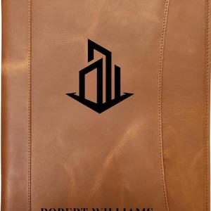 May include: A brown leather portfolio with a black architectural logo and the name "ROBERT WILLIAMS" printed at the bottom. The portfolio has a stitched border and a vertical seam on the right side.