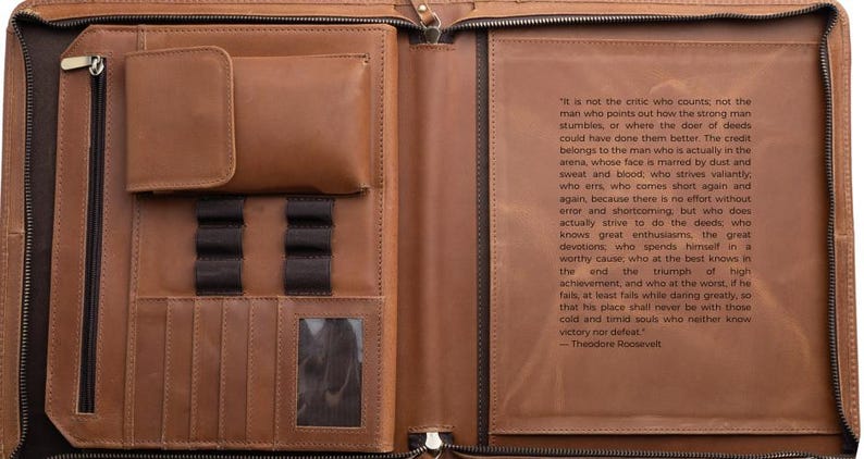 May include: Open leather portfolio with multiple pockets, pen holders, and a clear ID window. The interior features a quote by Theodore Roosevelt. The portfolio is brown with a zipper closure.