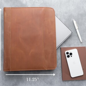 May include: A brown leather portfolio, 13.5 inches tall and 11.25 inches wide, with a zipper closure. A silver laptop, white pen, a brown notebook, and a white smartphone are next to the portfolio on a gray surface.