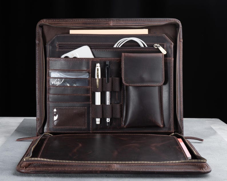 May include: Open brown leather portfolio with multiple compartments, including card slots, pen holders, and a small pocket. The portfolio is open, revealing a smartphone, pens, and a notepad. The zipper is visible around the edges.