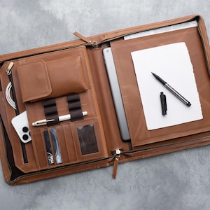 May include: Open brown leather portfolio with multiple compartments. Includes a phone, tablet, pen, notepad, and card slots. The portfolio is zippered and has a classic, professional design.
