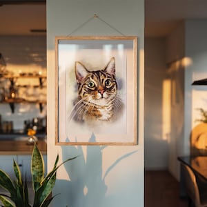 May include: A framed watercolor print of a tabby cat with brown and black stripes and large, expressive eyes. The artwork is displayed in a light-colored wooden frame, hanging on a pale green wall. The cat's portrait is the focal point.