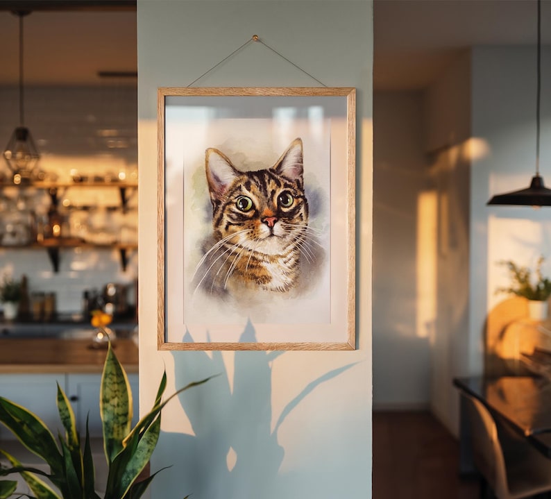 May include: A framed watercolor portrait of a tabby cat with brown and black stripes, large yellow eyes, and long white whiskers. The artwork is displayed in a light-colored wooden frame, hanging on a light blue wall.