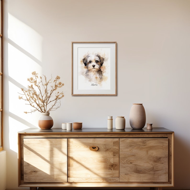 May include: A framed watercolor portrait of a small dog with a soft, neutral color palette. The dog's name, "Marta," is written below the image. The artwork is displayed on a wall above a wooden cabinet with decorative items.