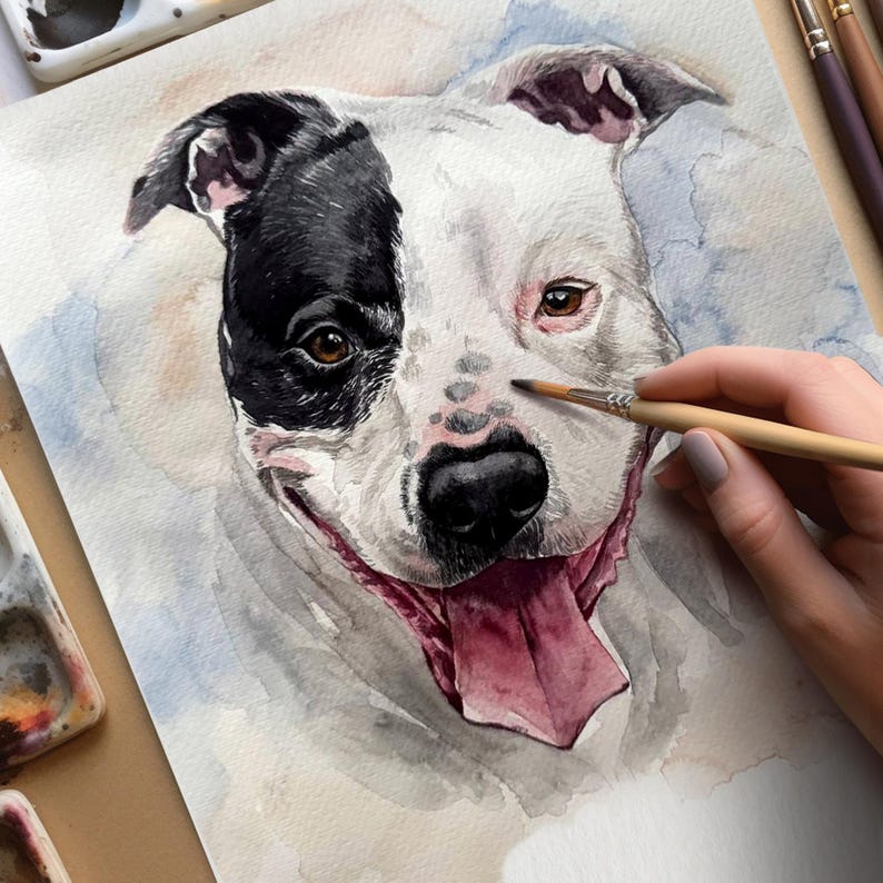 May include: Watercolor painting of a dog's face, featuring a black and white coat with brown eyes and a pink tongue. The artist's hand holds a paintbrush, adding details to the dog's nose. The artwork is on white paper with watercolor paint and brushes visible.
