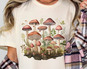 May include: A white t-shirt with a graphic of a cluster of mushrooms in various sizes and colors. The mushrooms are surrounded by green leaves and ferns.