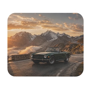 May include: A dark green vintage convertible sports car parked on an asphalt mountain road. The vehicle features a low profile, round headlights, and silver wheels. In the background, snow-capped mountain peaks are illuminated by a golden sunset.