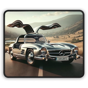 Może przedstawiać: Srebrny samochód sportowy Mercedes-Benz 300 SL Gullwing z lat 50. XX wieku z otwartymi drzwiami, zaparkowany na utwardzonej drodze z górami w tle. Samochód ma tablicę rejestracyjną z napisem "CARSARTS".