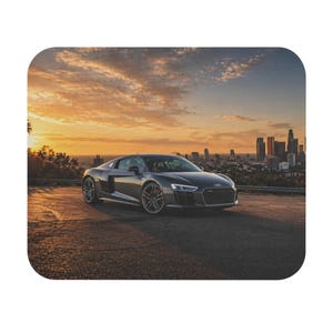 May include: A dark gray sports car is parked on a road, with a city skyline in the background. The sky is filled with orange and yellow hues from the setting sun. The car has a sleek design with black rims.