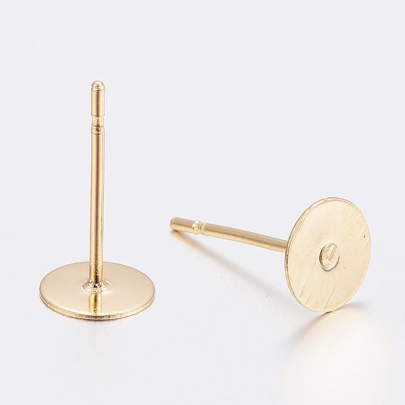 May include: Two gold-coloured earring posts with flat, round pads. One post is upright, showing the full length of the post and pad. The other post is lying down, showing the back of the pad and the post's details. These are jewellery making supplies.