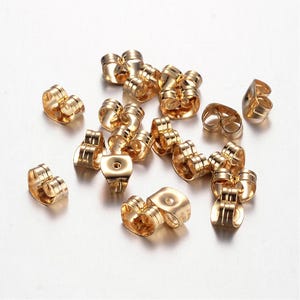 May include: A selection of gold-coloured earring backs scattered on a white surface. The earring backs are in various shapes, including butterfly and barrel styles. The metal has a shiny finish, reflecting light.