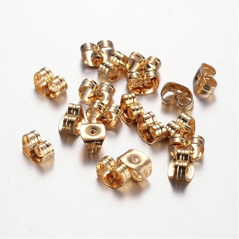 May include: A selection of gold-coloured earring backs scattered on a white surface. The earring backs are in various shapes, including butterfly and barrel styles. The metal has a shiny finish, reflecting light.