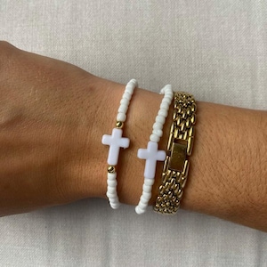 Puede incluir: Dos pulseras de cuentas blancas con un dije de cruz y un reloj dorado con una correa de eslabones. Las pulseras tienen cuentas blancas y una cruz blanca con detalles dorados. El reloj tiene una correa y un cierre dorados.