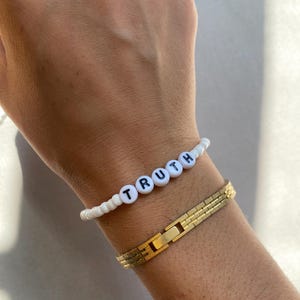 May include: A white beaded bracelet with the word "TRUTH" spelled out in black letters. A gold-colored, rectangular link bracelet is also visible.