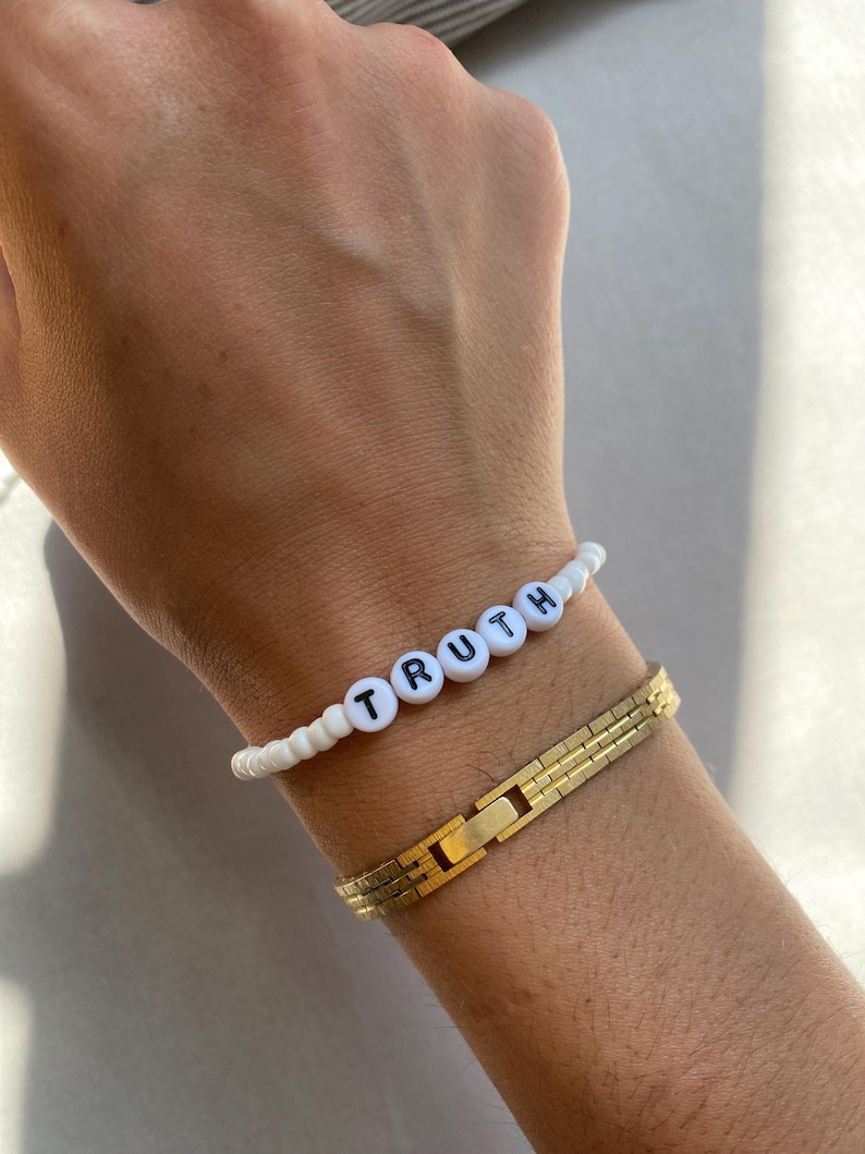 May include: A white beaded bracelet with the word "TRUTH" spelled out in black letters. A gold-colored, rectangular link bracelet is also visible.