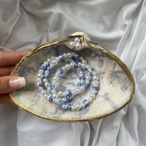 May include: A white seashell with a gold rim, decorated with a blue and white floral pattern, holds a white pearl necklace with blue floral accents.