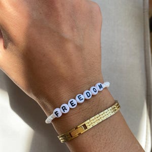 May include: A white beaded bracelet spelling "FREEDOM" in black letters. A gold-colored, rectangular link bracelet is worn below it. The bracelets are on a wrist.