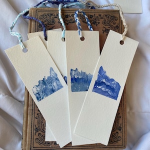 May include: Four white bookmarks with blue abstract watercolor designs. Each bookmark has a hole punched in the top and a white string attached.