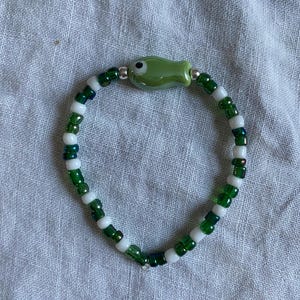 May include: A beaded bracelet featuring a green fish charm. The bracelet is made of small, round beads in shades of green and white. The fish charm is a light green color with a black eye and silver accents. The bracelet is shown on a light gray fabric.
