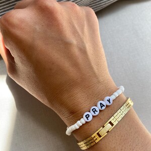 May include: Two bracelets on a wrist. One is white with black letter beads spelling "PRAY". The other is a gold-colored, rectangular link bracelet. The background is a neutral color.