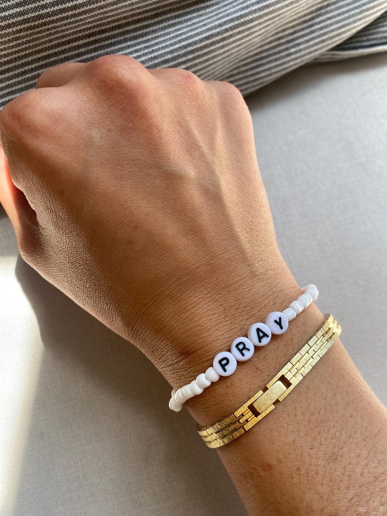 May include: Two bracelets on a wrist. One is white with black letter beads spelling "PRAY". The other is a gold-colored, rectangular link bracelet. The background is a neutral color.