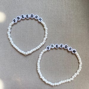 May include: Two white beaded bracelets with the word "FREEDOM" spelled out in black letters on white letter beads. The bracelets are on a neutral-colored surface.