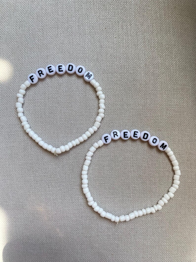 May include: Two white beaded bracelets with the word "FREEDOM" spelled out in black letters on white letter beads. The bracelets are on a neutral-colored surface.