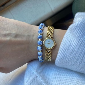 May include: A gold watch with a white face and black Roman numeral markings. The watch is on a gold metal band. A white and blue beaded bracelet is worn on the same wrist.