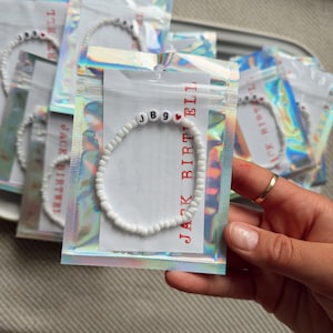 May include: White beaded bracelet with the letters "JB9" and a red heart charm. The bracelet is packaged in a clear, iridescent pouch with the text "JACK BIRTH ELI" printed in red. Several bracelets are visible.