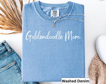Può includere: Una maglietta azzurra con la scritta bianca "Goldendoodle Mom". Una collana d'argento con un ciondolo poggia sulla maglietta. La maglietta è piegata, con un paio di jeans e una pianta secca visibili.