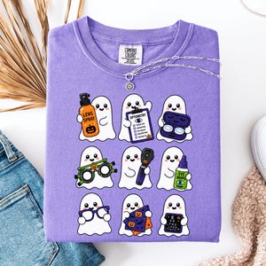 May include: A violet t-shirt featuring a graphic print of nine cartoon ghosts. Each ghost holds an optometry-related item, including lens spray, an eye chart, contact lens cases, trial frames, an ophthalmoscope, eye drops, glasses, and a cloth.