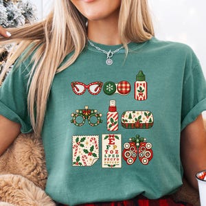 Könnte beinhalten: Ein salbeigrünes T-Shirt mit einem Grafikdruck verschiedener Augenpflegeartikel im Weihnachtsthema, darunter eine rote Brille, ein Kontaktlinsenbehälter, ein Fläschchen, eine festliche Sehtafel und ein Phoropter, verziert mit Stechpalmen und Karomustern.