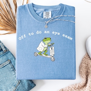 Puede incluir: Camiseta azul claro con el texto "Off to do an eye exam". La camiseta presenta una rana de dibujos animados con bata de laboratorio montada en un scooter y sosteniendo una tabla optométrica. Un collar plateado está sobre la camiseta.