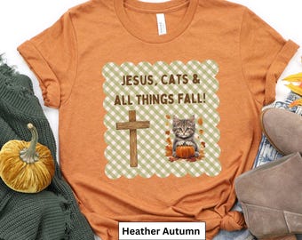 May include: Heather autumn-colored t-shirt with the text "JESUS, CATS & ALL THINGS FALL!" featuring a cross and a cat holding a pumpkin. The shirt has a round neck and short sleeves. A pumpkin and a green blanket are in the background.