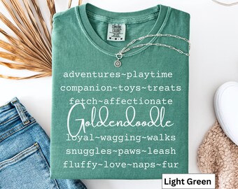 Può includere: T-shirt verde chiaro con la parola "Goldendoodle" in corsivo. La maglietta presenta un elenco di parole associate ai Goldendoodle, come "avventure", "tempo di gioco" e "soffice". Una collana d'argento è drappeggiata sulla maglietta.
