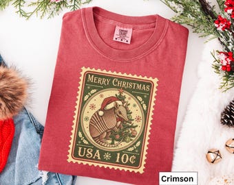 May include: Crimson-colored t-shirt featuring a Christmas-themed stamp design. The stamp depicts an armadillo wearing a Santa hat next to a decorated Christmas tree. The stamp reads "Merry Christmas USA 10¢".