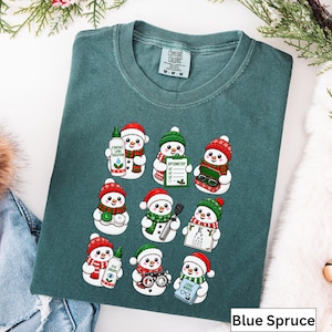 May include: A blue spruce coloured t-shirt featuring a graphic print of nine cartoon snowmen. Each snowman is depicted with optometry-themed items including contact lens solution, an eye chart, glasses, eye drops, and a lens cleaning kit.