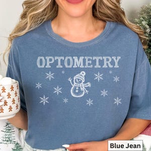 May include: A blue jean colored t-shirt featuring the white text OPTOMETRY centered above a graphic of a snowman wearing a hat and scarf, surrounded by various white snowflake illustrations.