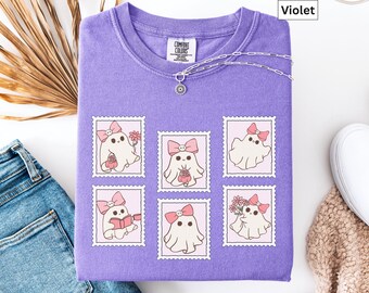 May include: A violet Comfort Colors sweatshirt featuring a design of six ghost stamps. Each stamp shows a cute ghost with a pink bow, holding flowers, a book, or a basket. A silver necklace with a small pendant is on the shirt.