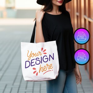 May include: A white tote bag with the text "Your DESIGN here" in purple and orange lettering. The bag is being held by a person wearing a black shirt and blue jeans.