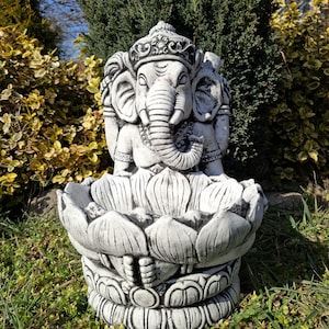 Puede incluir: Una estatua de hormigón blanco del dios hindú Ganesha, sentado sobre una base de flor de loto. La estatua está detallada con tallas intrincadas y tiene un diseño tradicional.